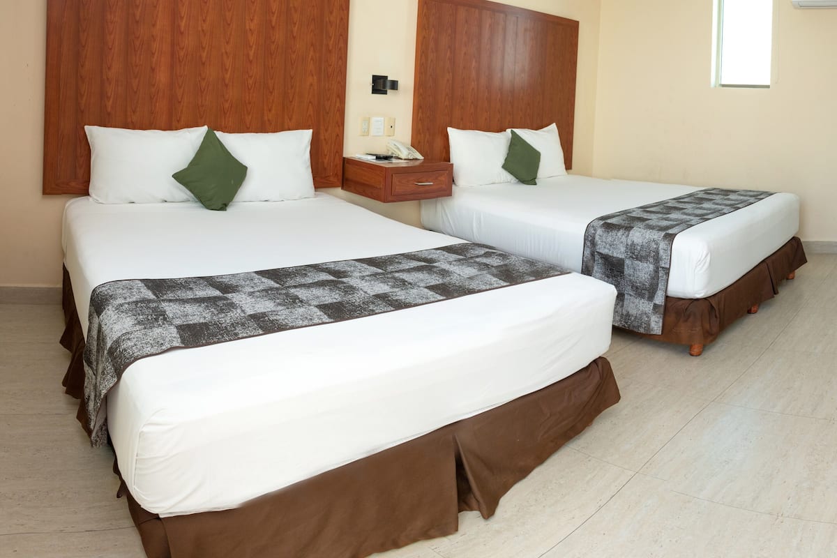 Dos camas dobles con cabeceros de madera, ropa de cama blanca y cubrelechos marrones en una habitación de hotel con piso de baldosas y una ventana con persianas.