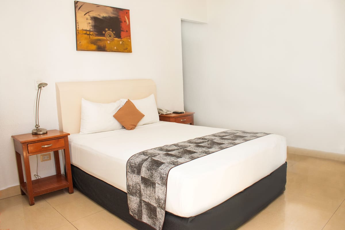 Una cama bien hecha con un edredón estampado, una lámpara de noche y una mesita de madera.