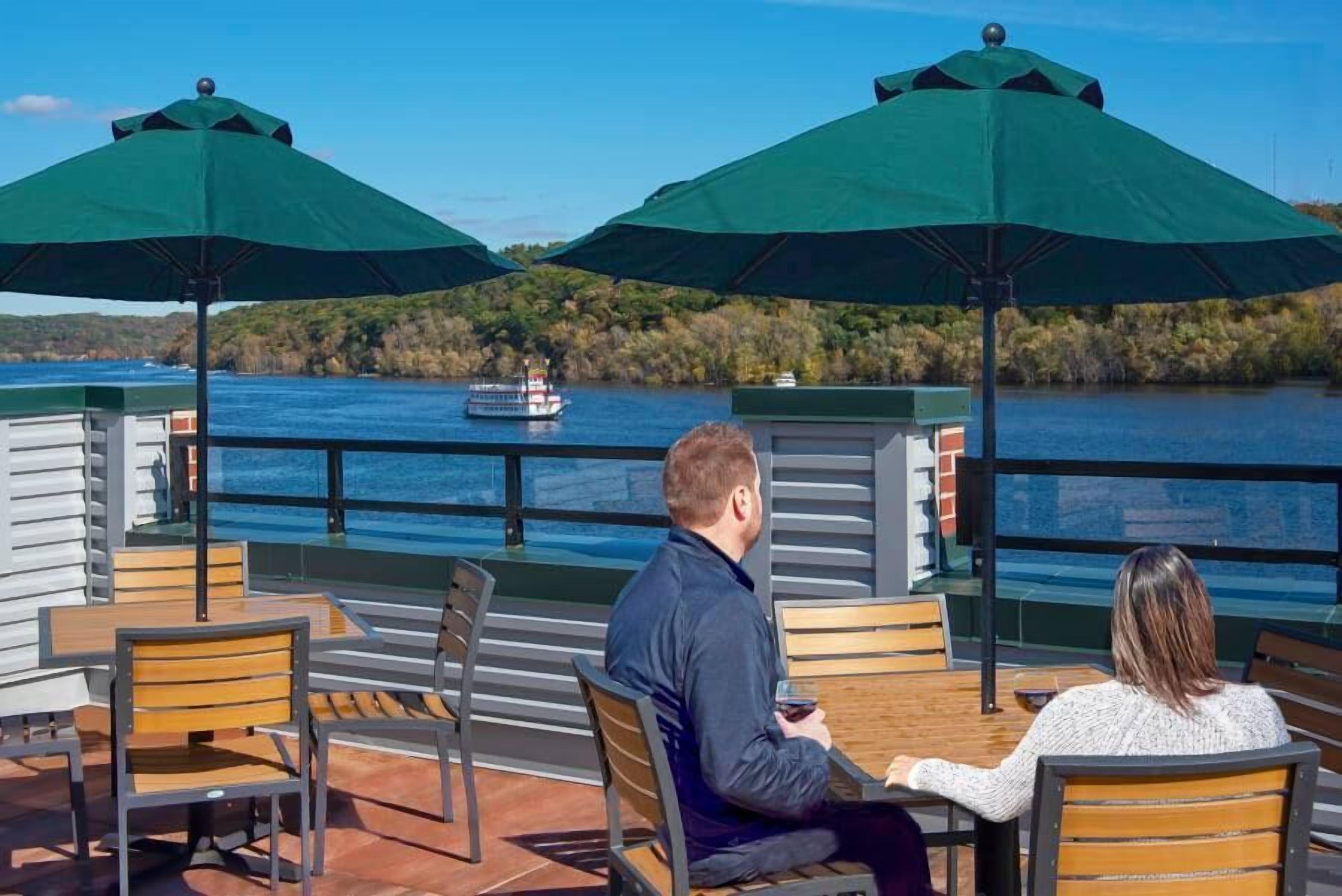 A terrace with wooden tables and chairs, green umbrellas, and a view of a boat on the water.