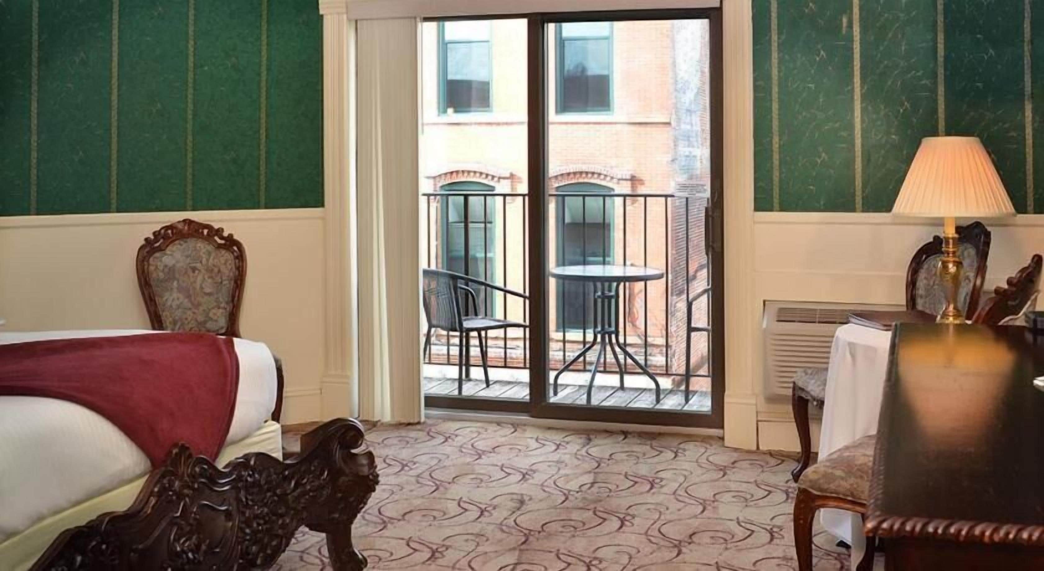 Habitación de hotel con cama, escritorio con lámpara, silla y balcón con mesa y sillas.