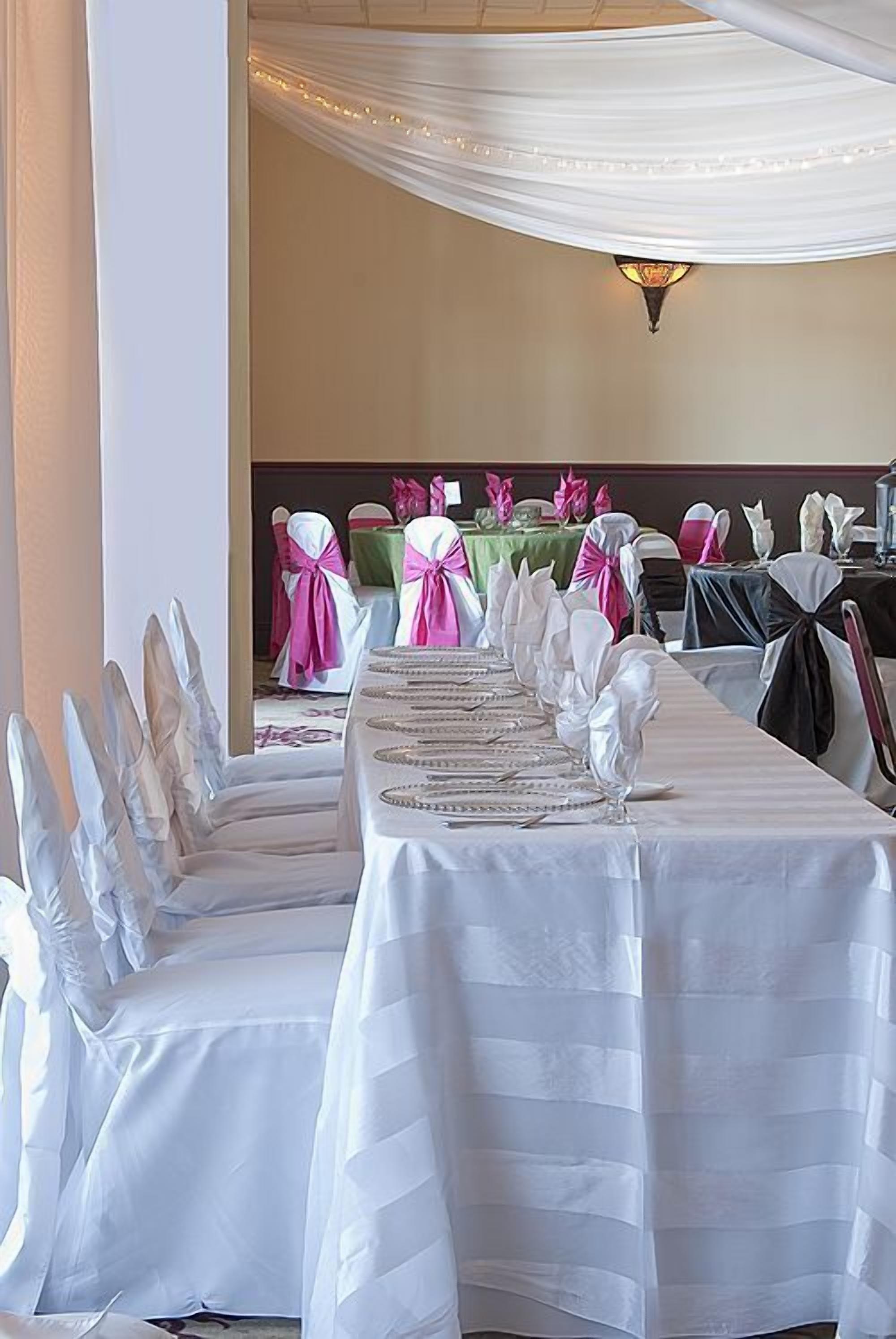 A banquet hall with white tablecloths, chairs with white and pink sashes, and a chandelier.