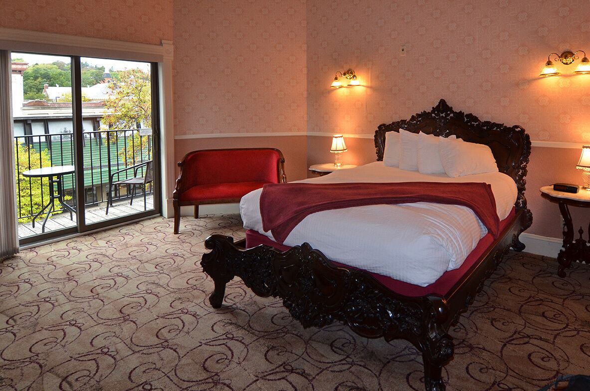 Habitación de hotel con una cama grande, una silla roja, un balcón con mesa y sillas, y un perro pequeño en el suelo.