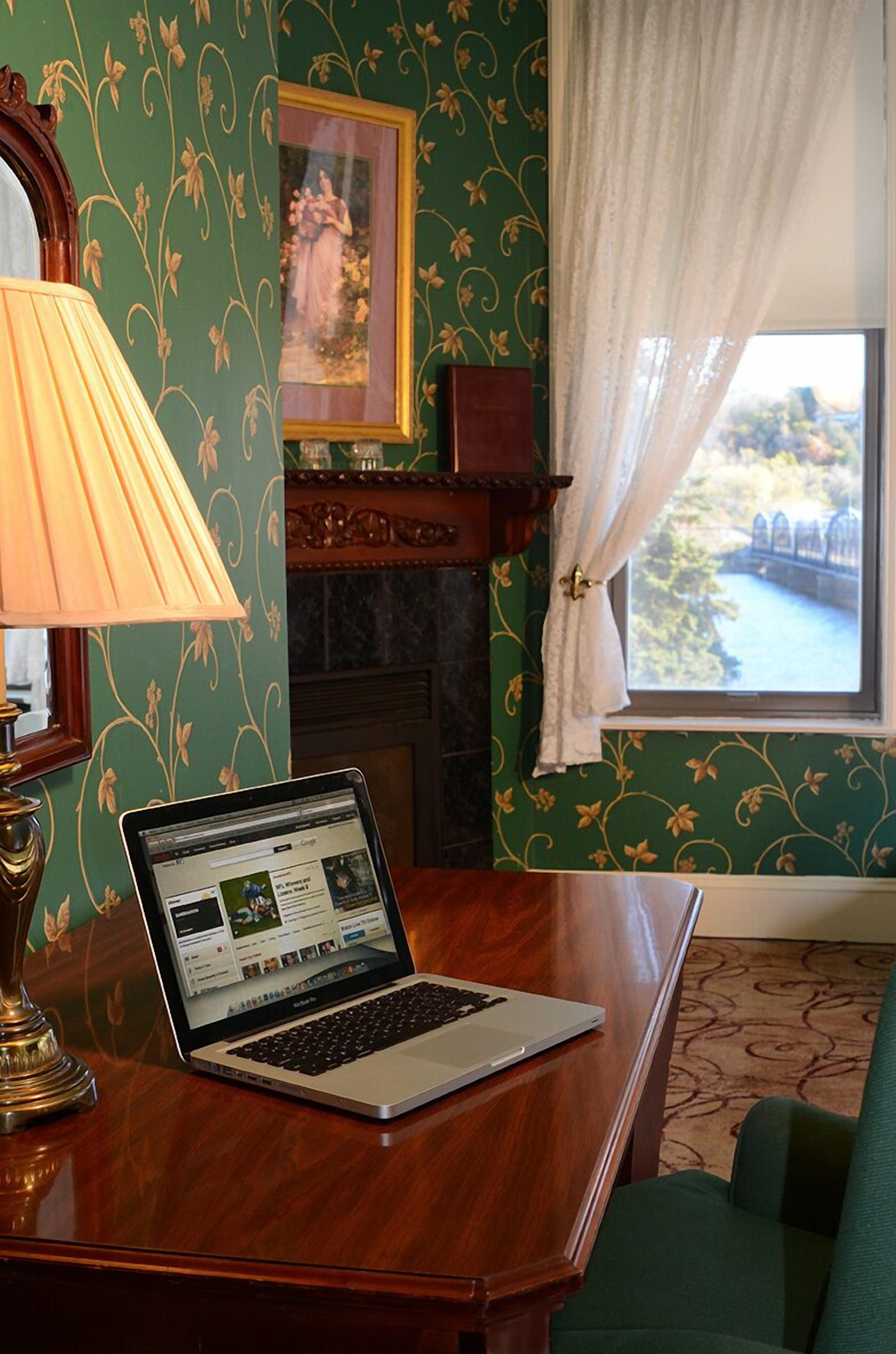 A room with a laptop on a wooden table, a lamp, a fireplace, a green patterned wall, a framed picture, and a window with a view of a bridge.
