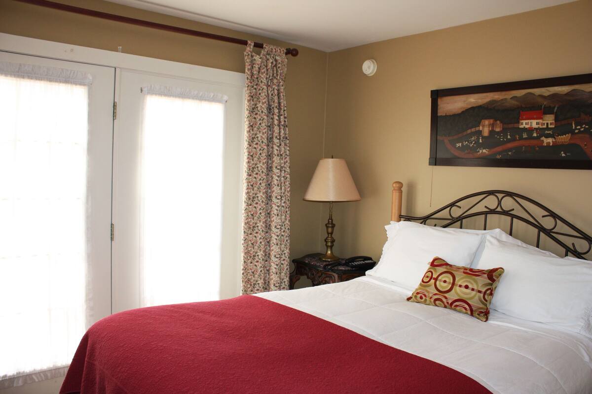 A bedroom with a bed, a nightstand, a lamp, a painting on the wall, and a window with curtains.