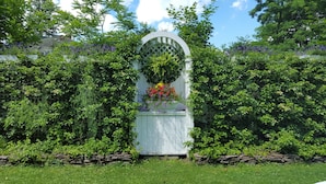 A garden with a white arbor surrounded by lush greenery and flowering plants. - The White House Inn (Cooperstown)