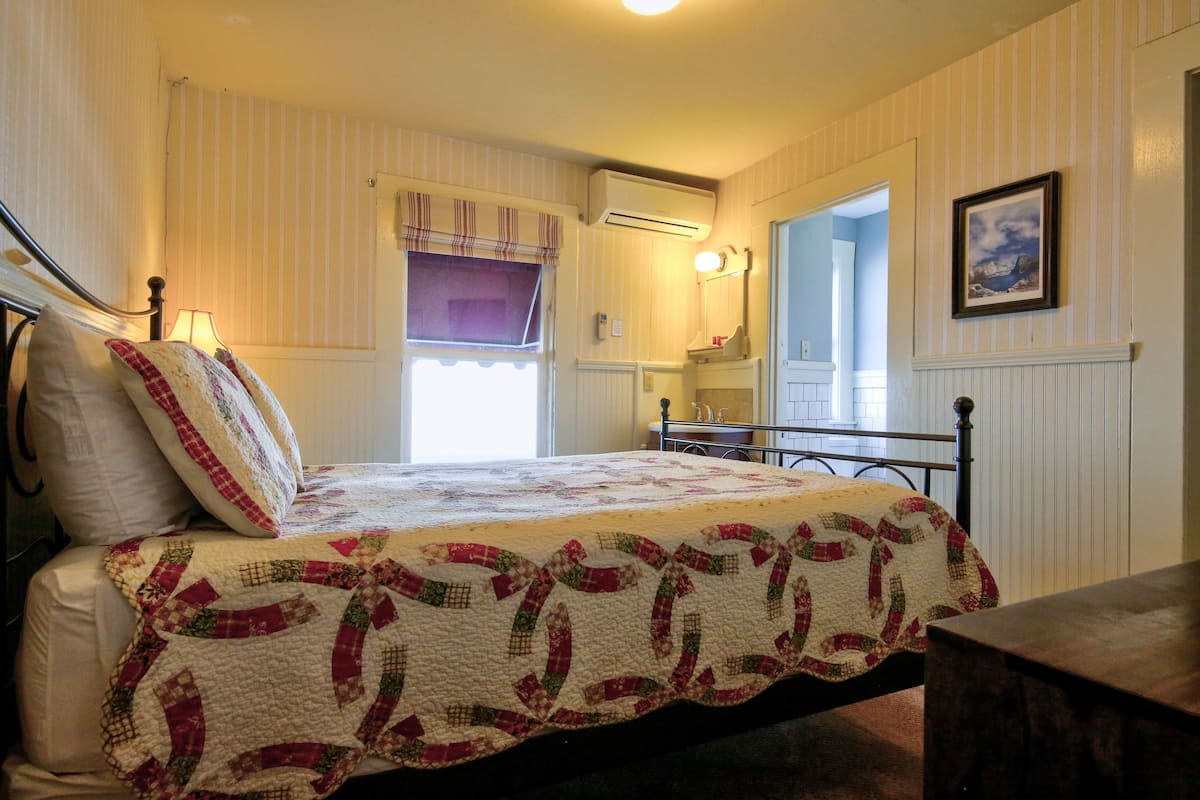 Une chambre à coucher comprenant un lit, une table de chevet, une fenêtre avec des rideaux, un climatiseur fixé au mur et un tableau encadré accroché au mur.