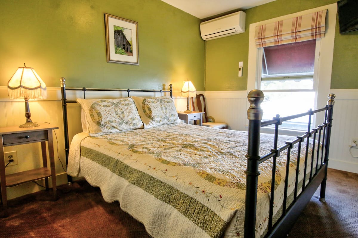 Une chambre à coucher avec un lit, deux tables de chevet avec des lampes, un climatiseur fixé au mur et une fenêtre avec un rideau.
