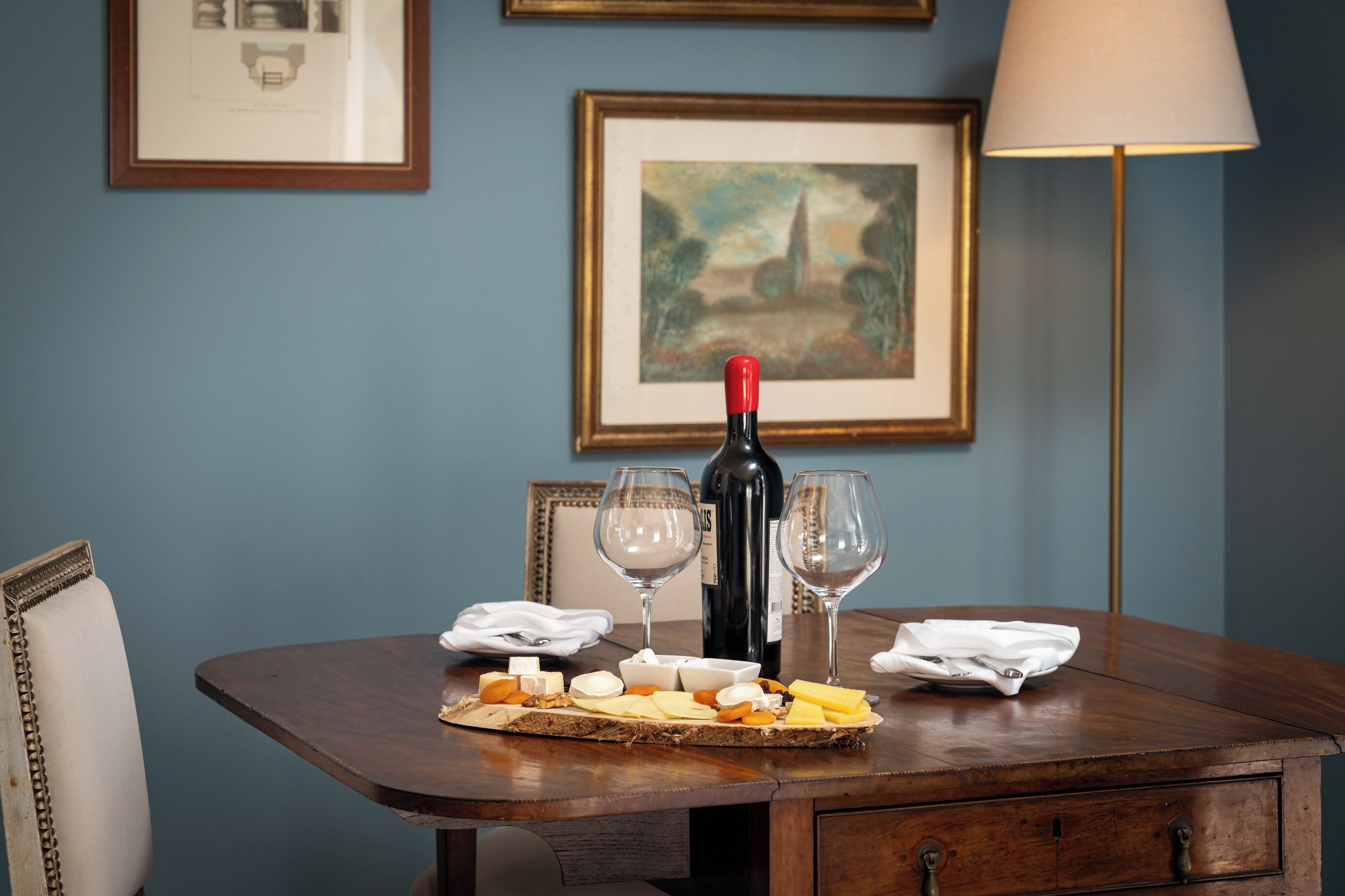 Mesa de jantar com tábua de queijos, garrafa de vinho e copos, em frente a uma parede azul com quadros.