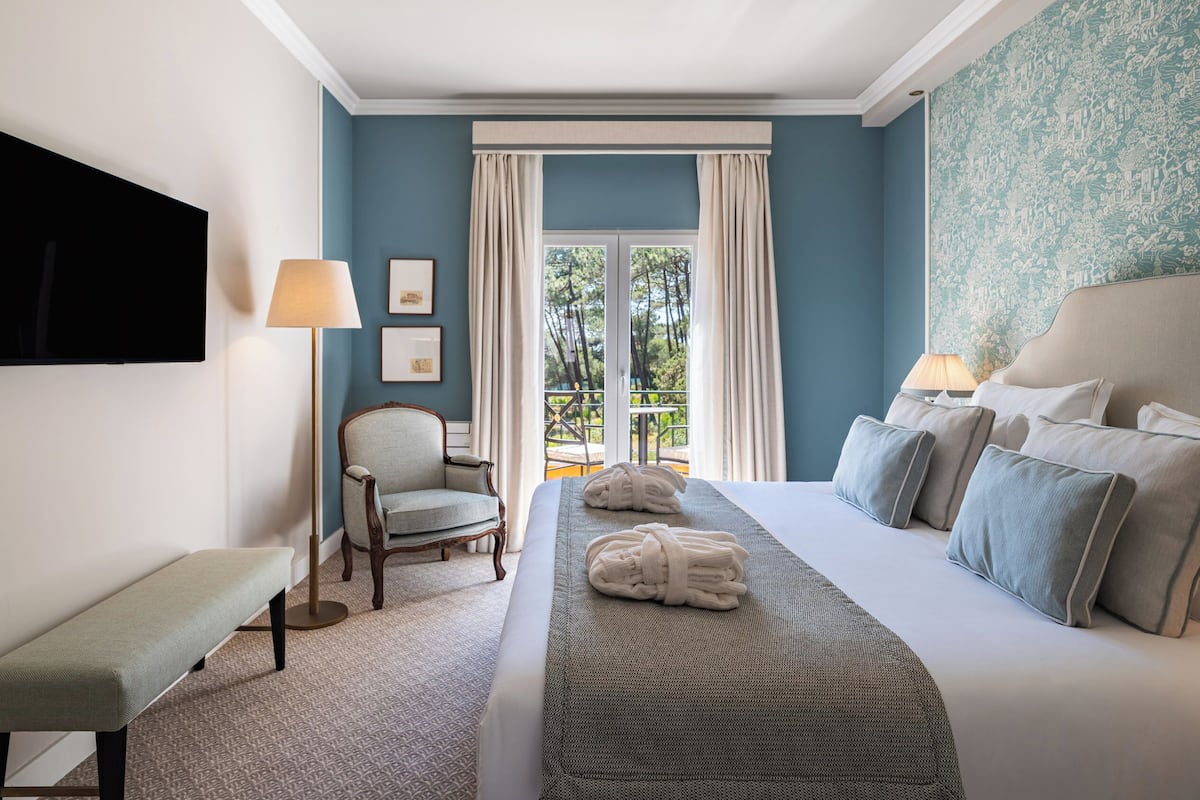 A hotel room with a large bed, a bench, a chair, a TV, and a balcony with curtains.