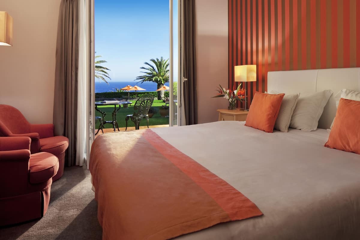 A hotel room with a large bed, a red armchair, a nightstand with a lamp, and a view of the ocean and outdoor seating area.
