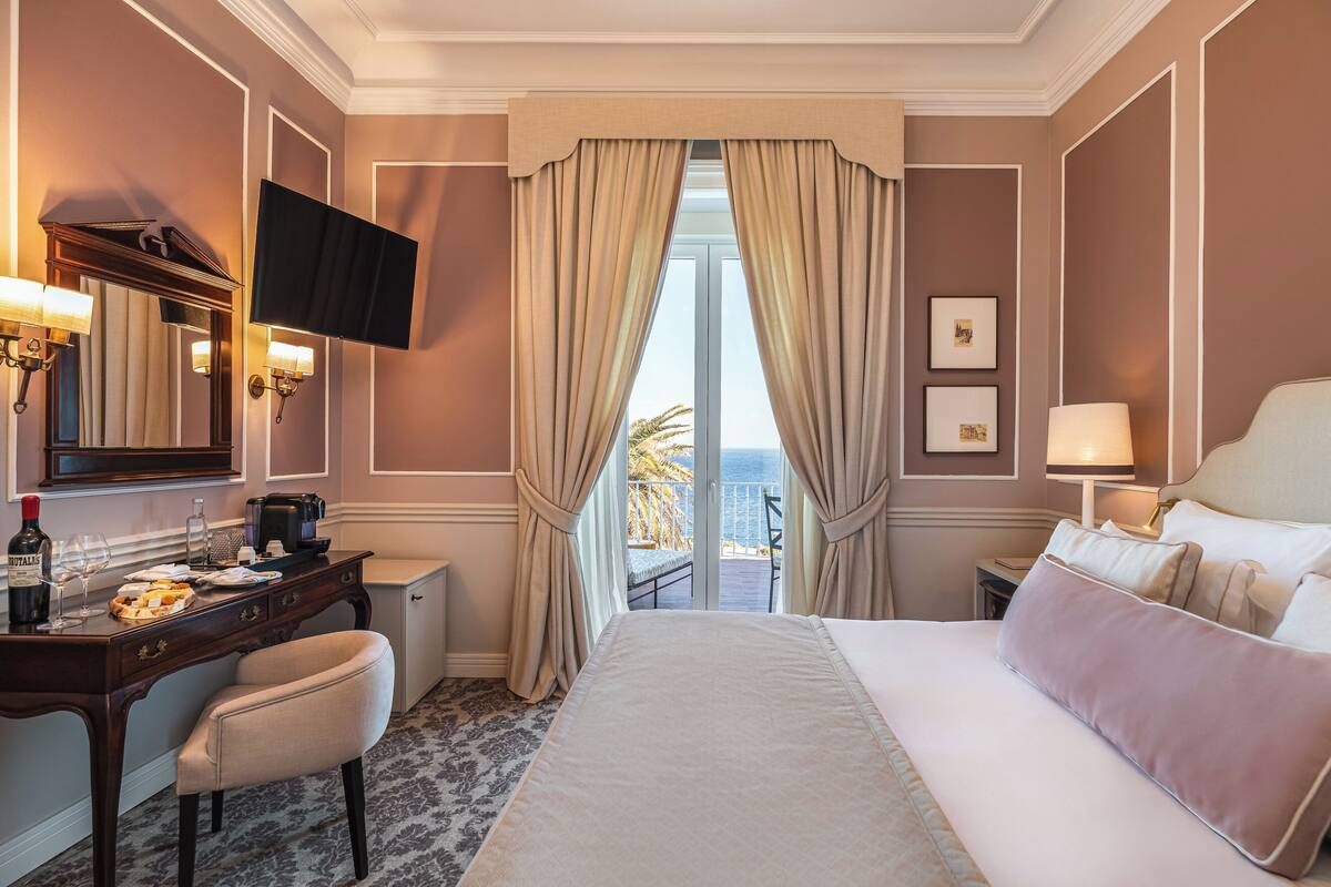 A hotel room with a large bed, a desk with a coffee maker, a TV mounted on the wall, and a view of the ocean through the window.