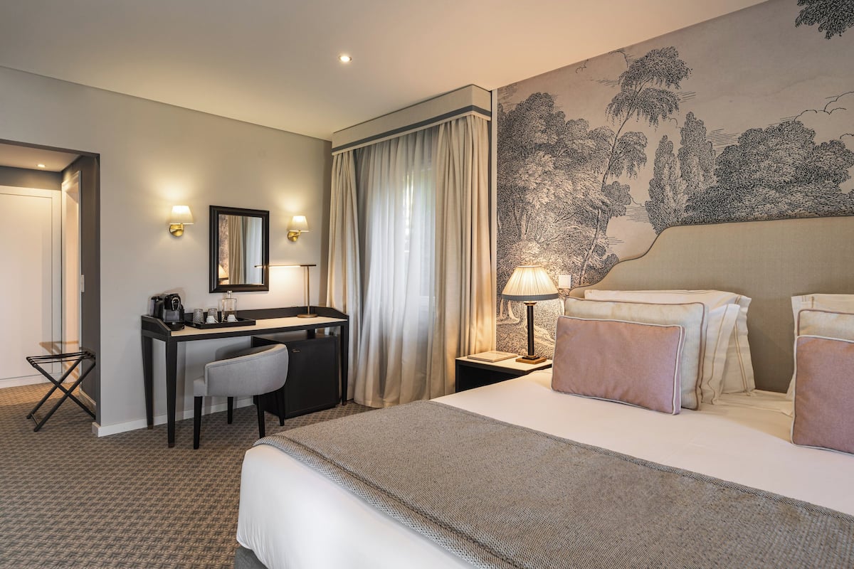 A hotel room with a bed, a desk, a chair, a lamp, a nightstand, and a wall with a landscape painting.