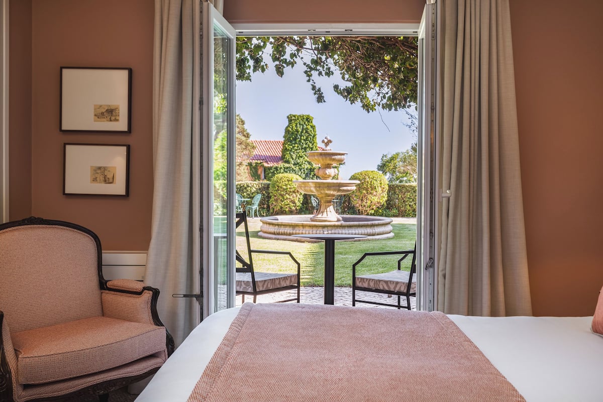 A hotel room with a bed, a chair, a table, and a view of a garden with a fountain.