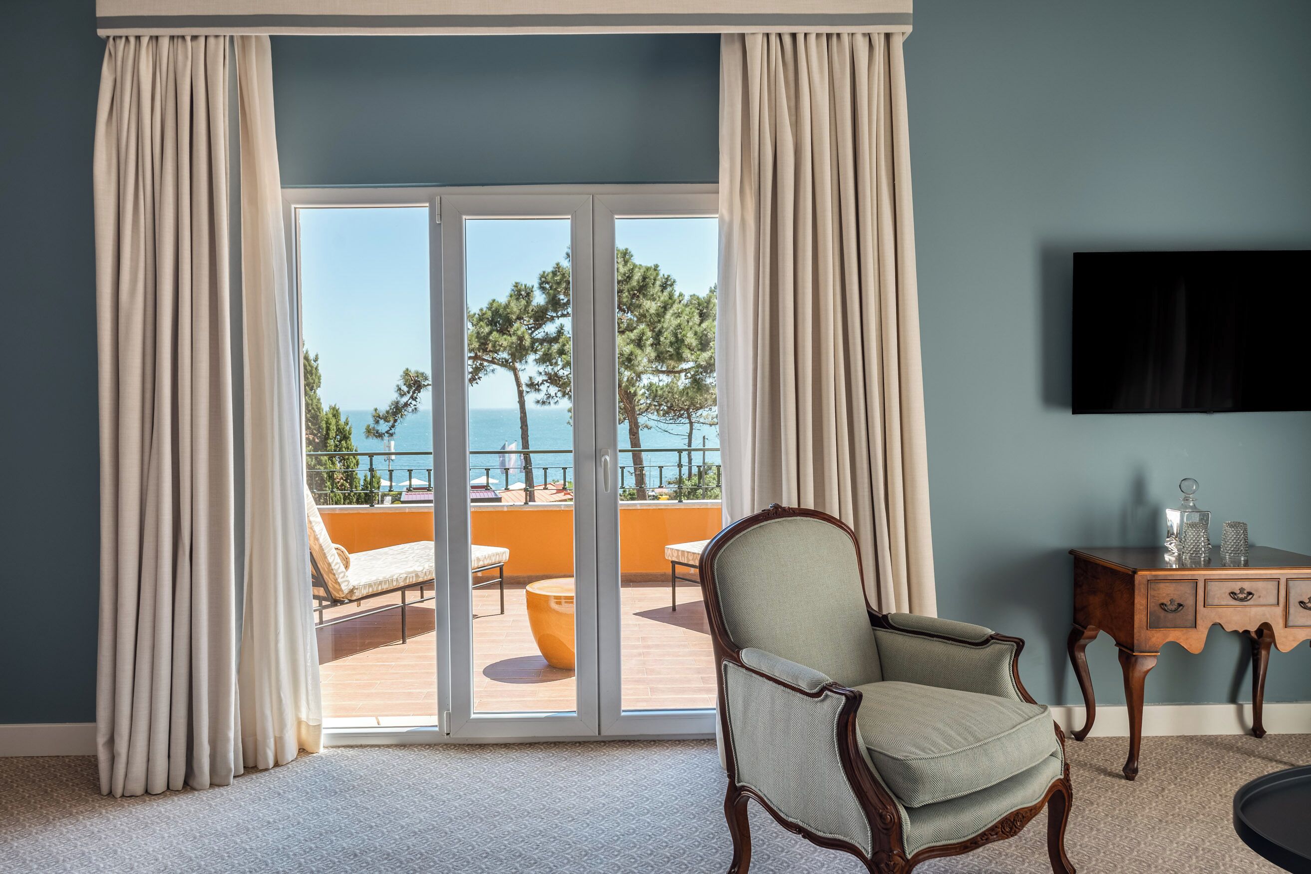 A room with a view of a balcony, a chair, a small table, and a flat-screen TV mounted on the wall.