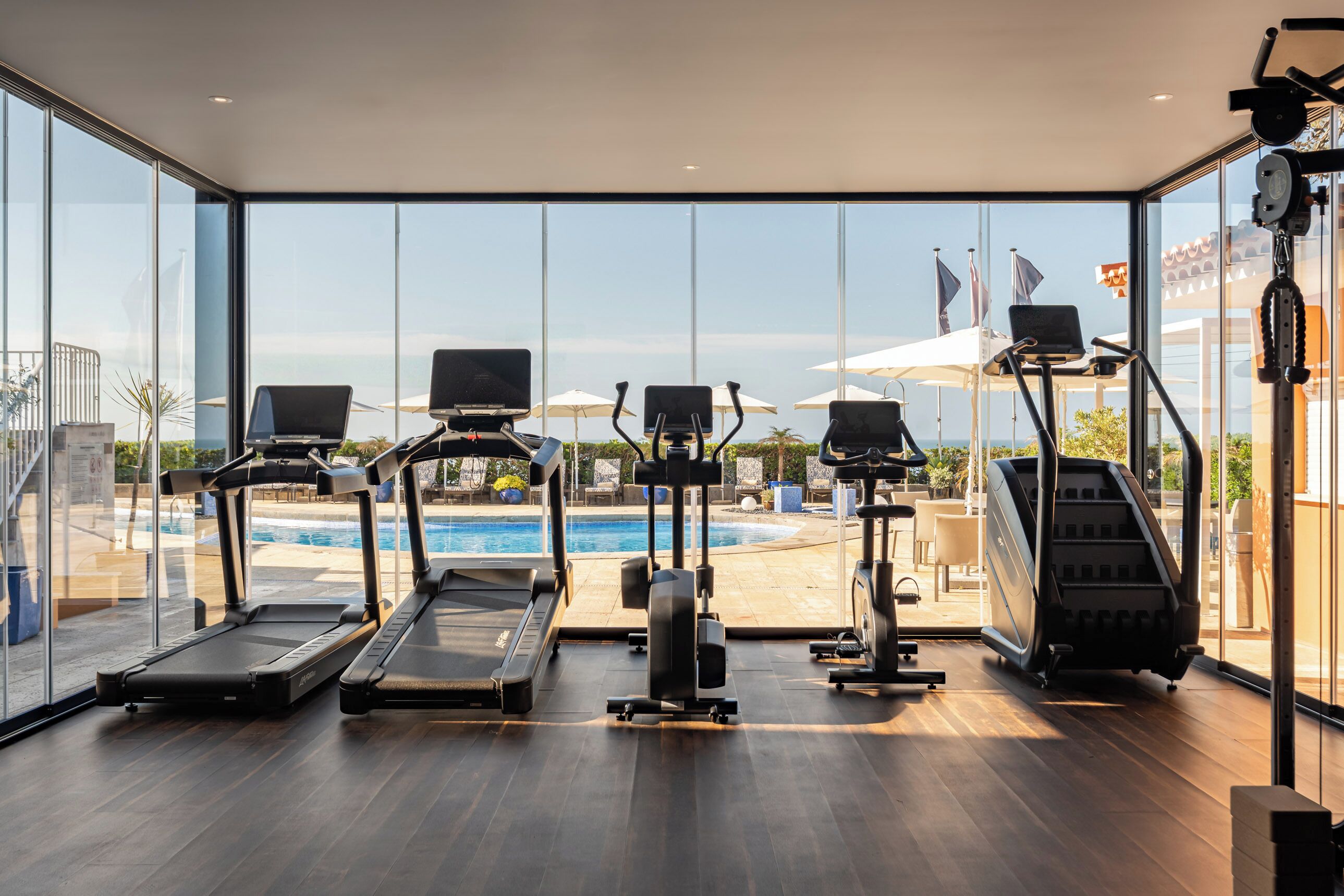 A modern gym with treadmills, elliptical machines, and a rowing machine, overlooking a pool and outdoor area.