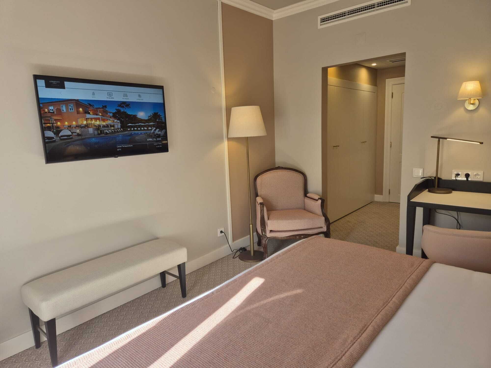 A hotel room with a bed, a desk, a chair, a bench, a TV mounted on the wall, and a lamp.