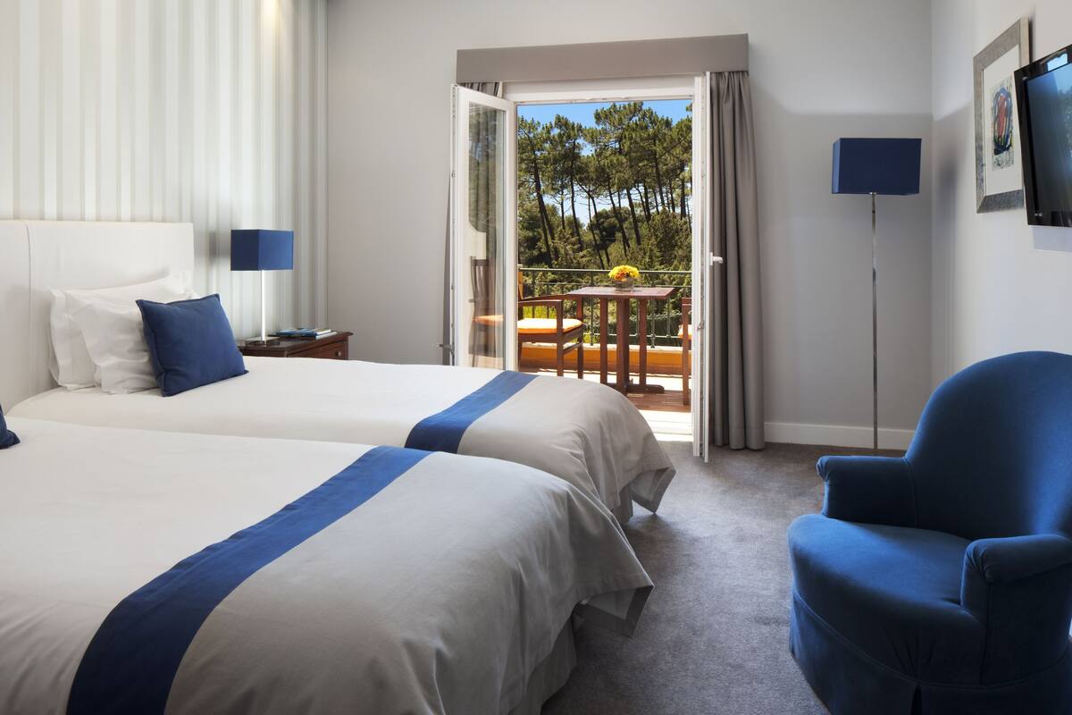A hotel room with two beds, a chair, a table with a vase, a TV, and a balcony with a view of trees.