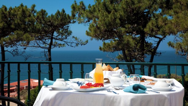 A table set with breakfast items, including fruit, bread, and a glass of orange juice, overlooking a scenic view of the sea and trees. - Longevity Senses - Cascais Boutique Hotel - Adults Only (Cascais)