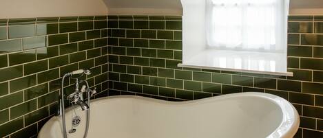 Un baño con una bañera independiente, paredes cubiertas de azulejos verdes y una ventana con cortinas blancas.