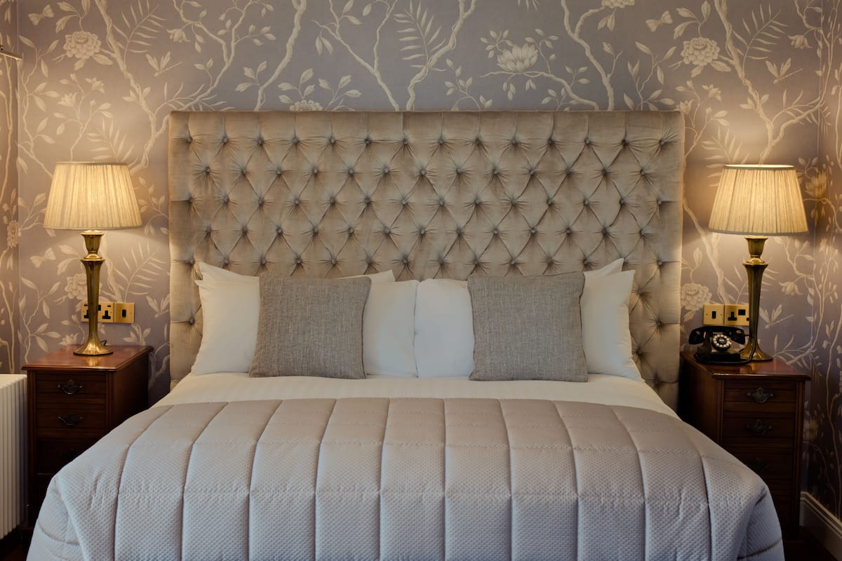 A neatly made bed with a tufted headboard, two bedside lamps, and a floral wallpaper pattern.