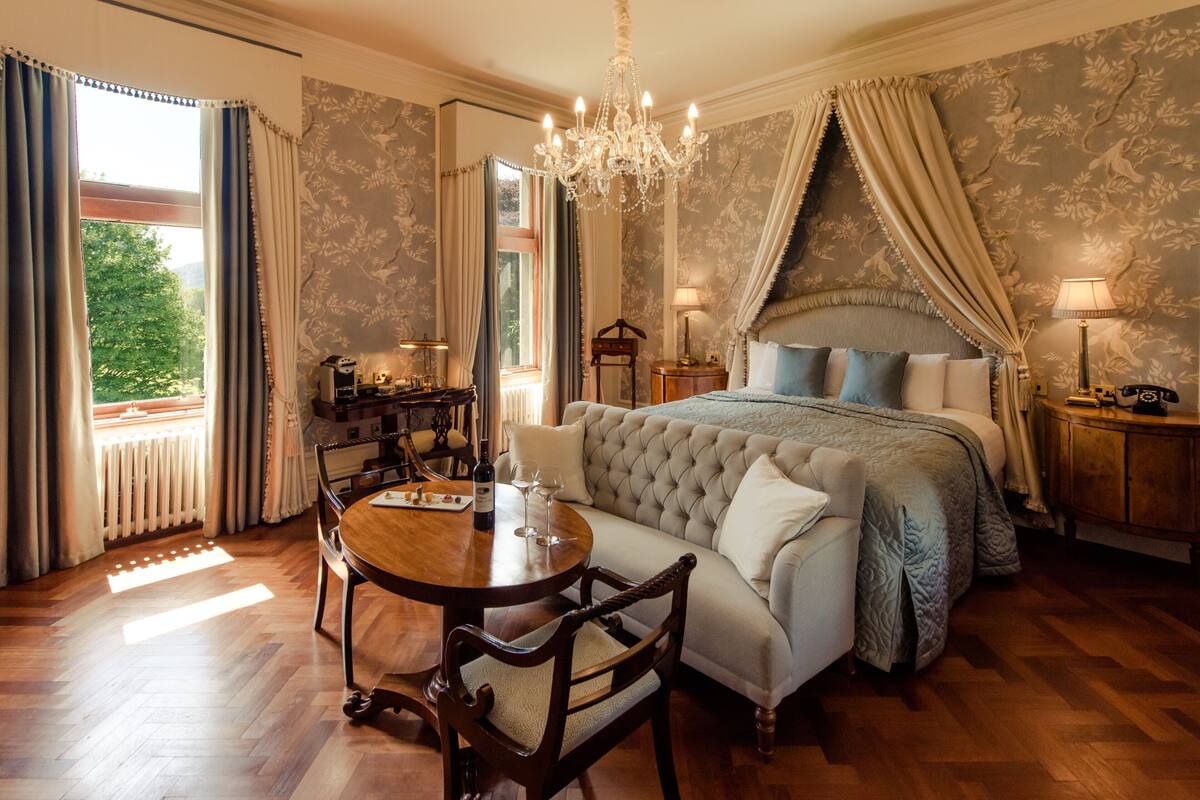 A bedroom with a large bed, a chandelier, a dining table with chairs, and a view of the outdoors.