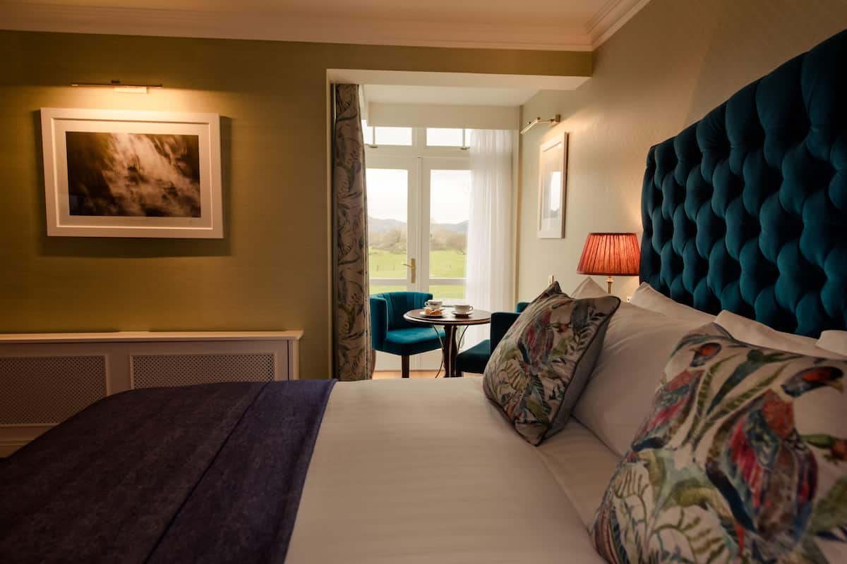 A hotel room with a large bed, a view of the outdoors, a small table with a lamp, and a framed picture on the wall.