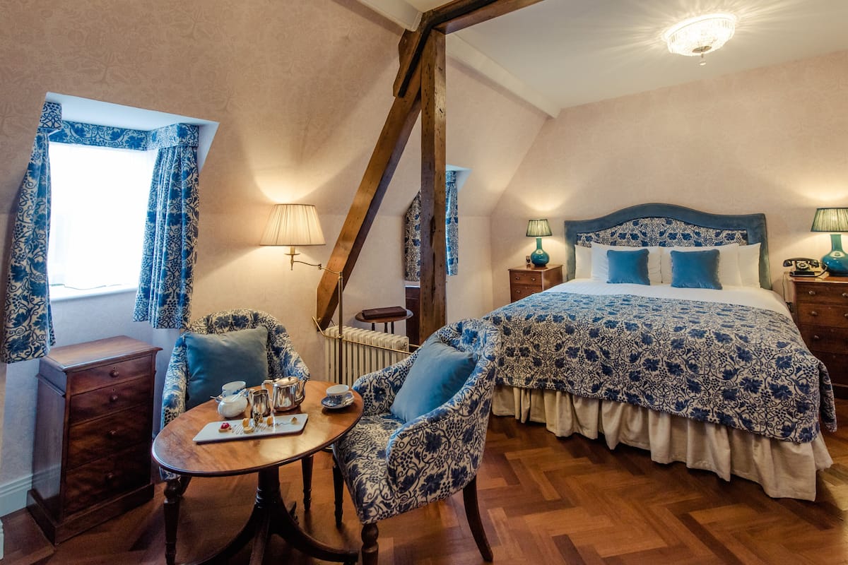 A bedroom with a large bed, a wooden chair, a table with a tray, and a window with curtains.