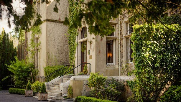 Un edificio histórico con escalones de piedra que conducen a una puerta, rodeado de abundante vegetación y un jardín bien cuidado.