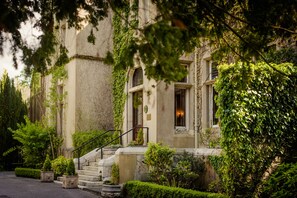 A historic building with stone steps leading to a door, surrounded by lush greenery and a well-maintained garden.