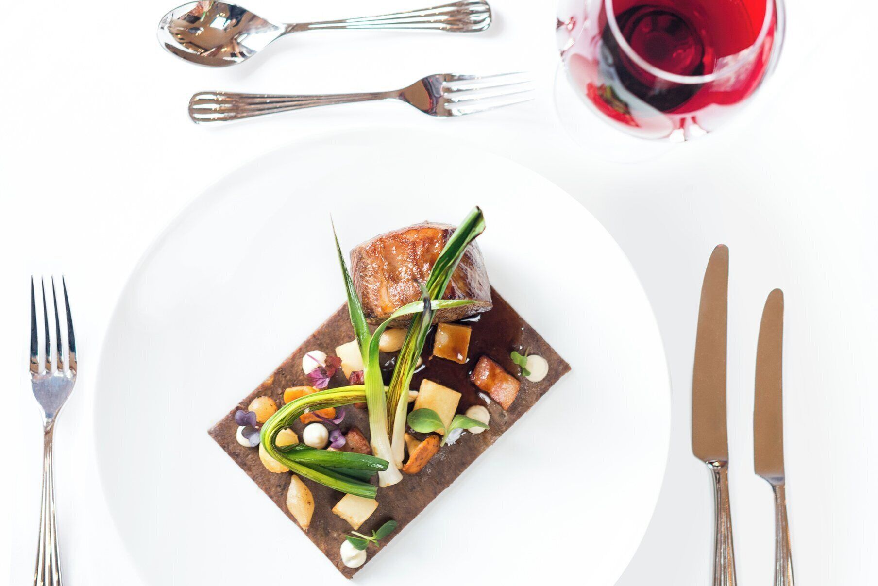A white plate with a square-shaped dish containing a piece of meat, vegetables, and a sauce, accompanied by cutlery and a glass of red wine.
