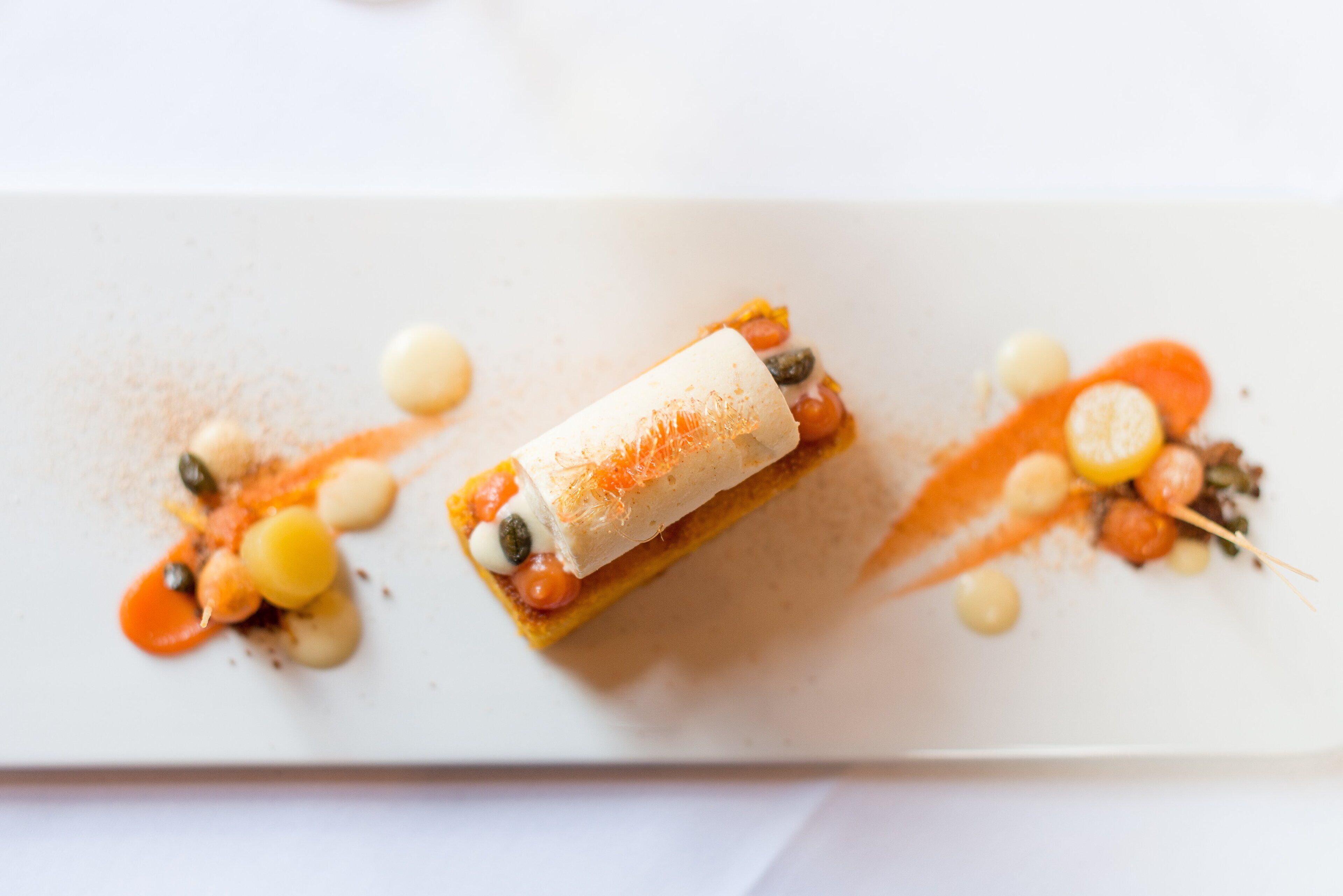 A white rectangular plate with a rolled pastry, small round garnishes, and a drizzle of sauce.
