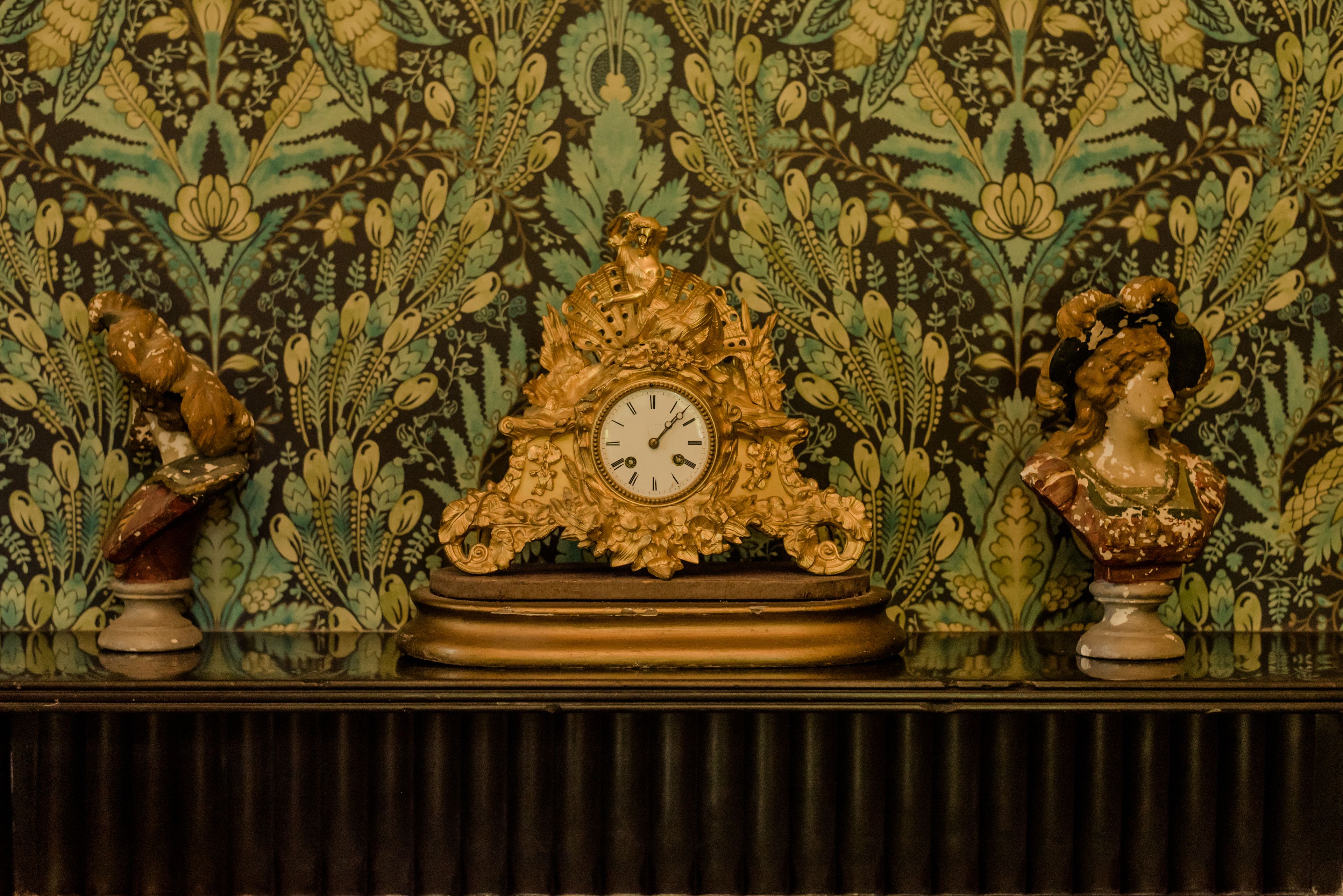 A decorative clock with ornate designs and two busts on either side, set against a patterned wallpaper background.