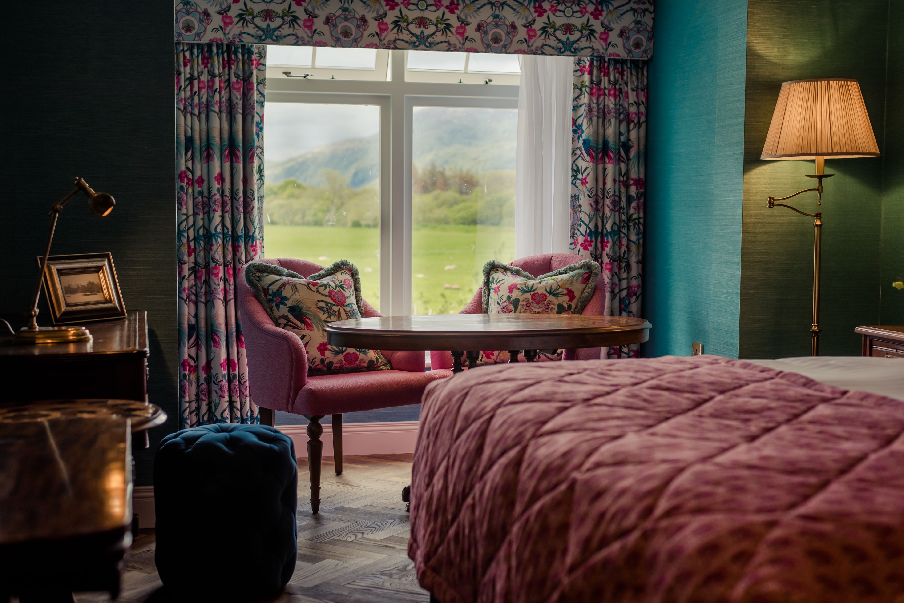 Una camera da letto con un letto grande, un tavolo rotondo, due sedie, una lampada e vista su un paesaggio verde attraverso una finestra.