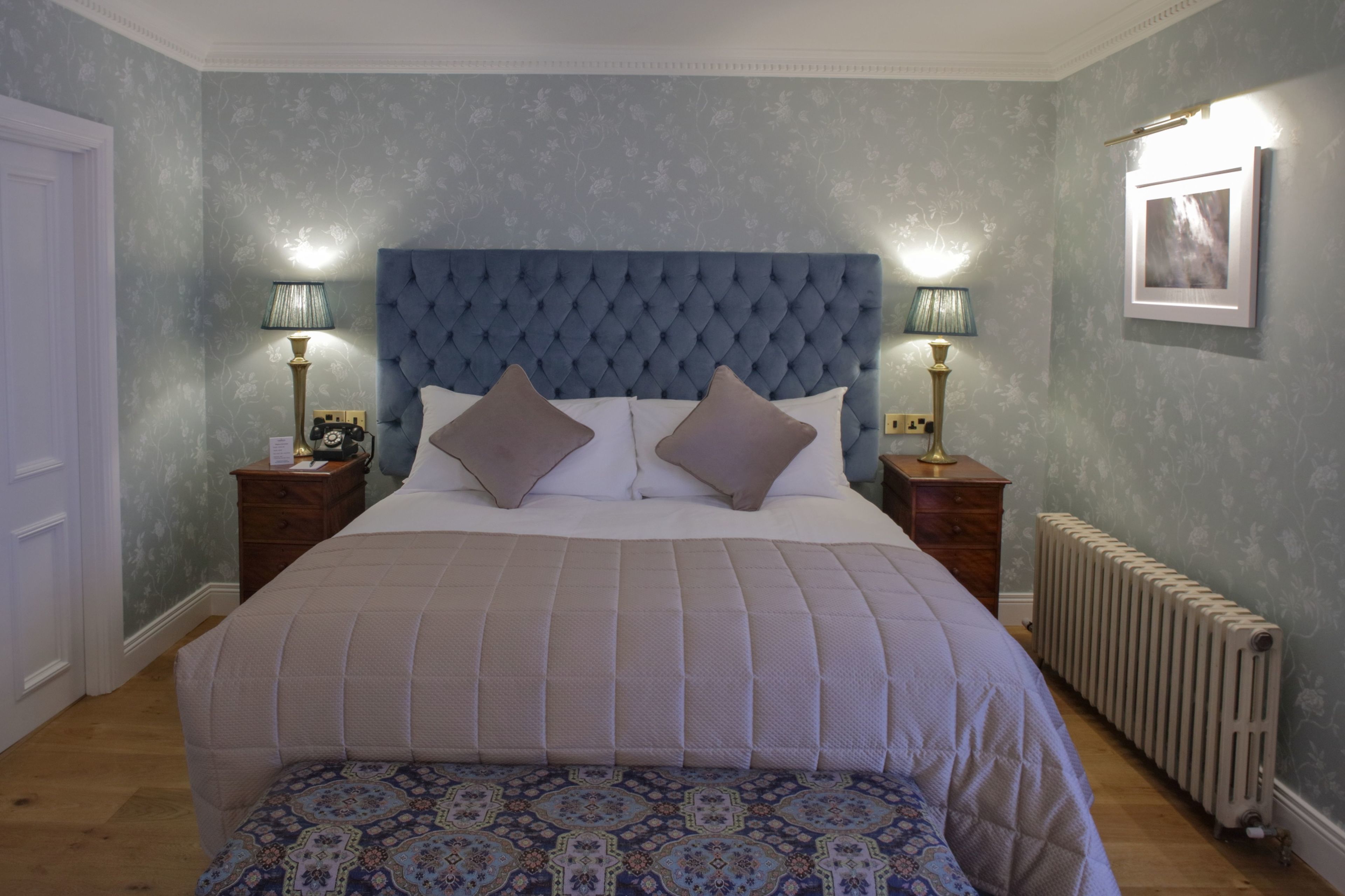 A bedroom with a tufted headboard, two bedside tables with lamps, a patterned bedspread, and a radiator.