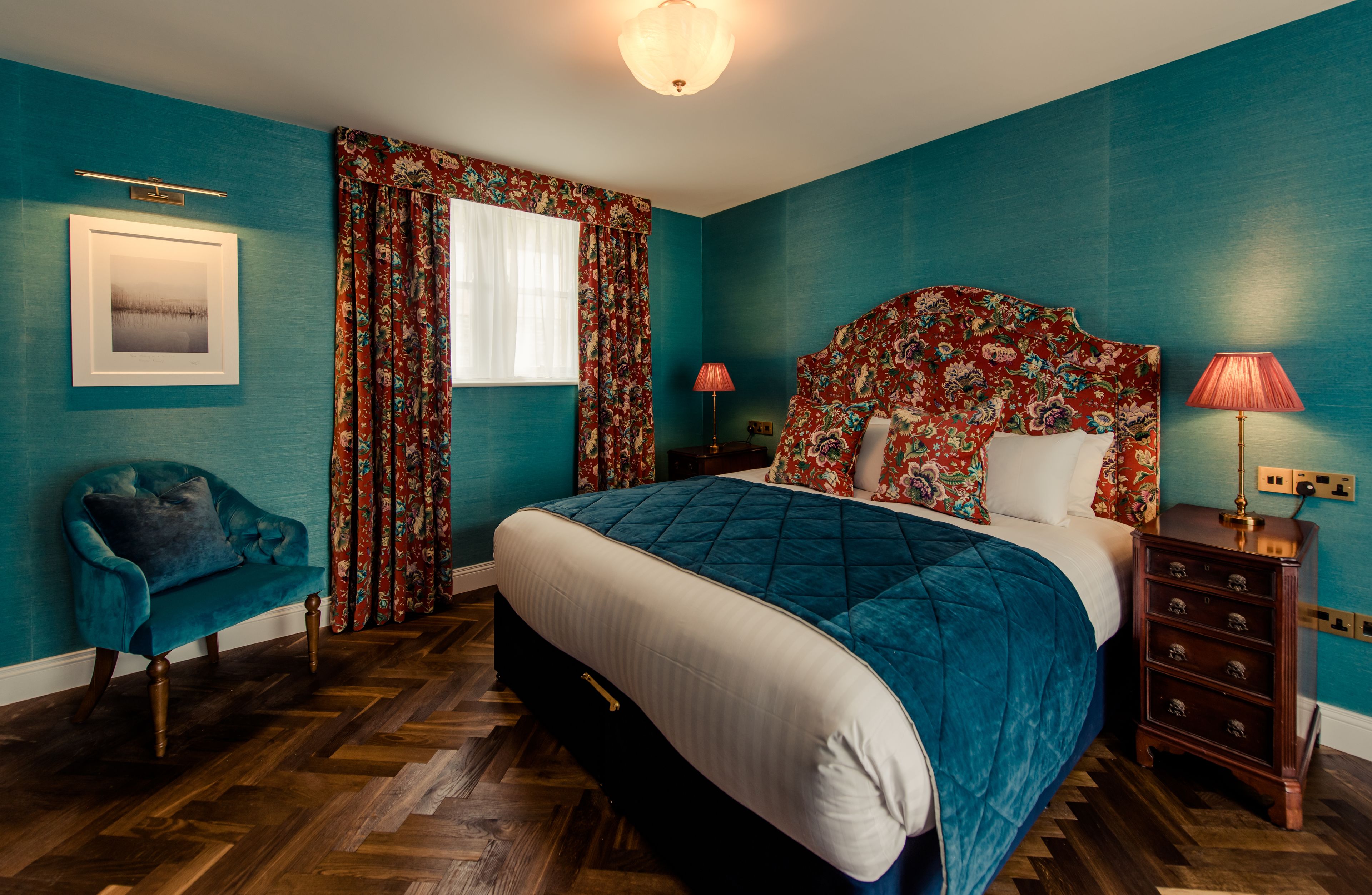 Quarto com paredes verde-água, cabeceira estampada com flores, cama com colcha, mesa de cabeceira de madeira, poltrona azul e janela com cortinas.