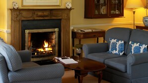 Una sala de estar con chimenea, dos sillones, un sofá, un aparador y una mesa con un libro y una copa de vino.