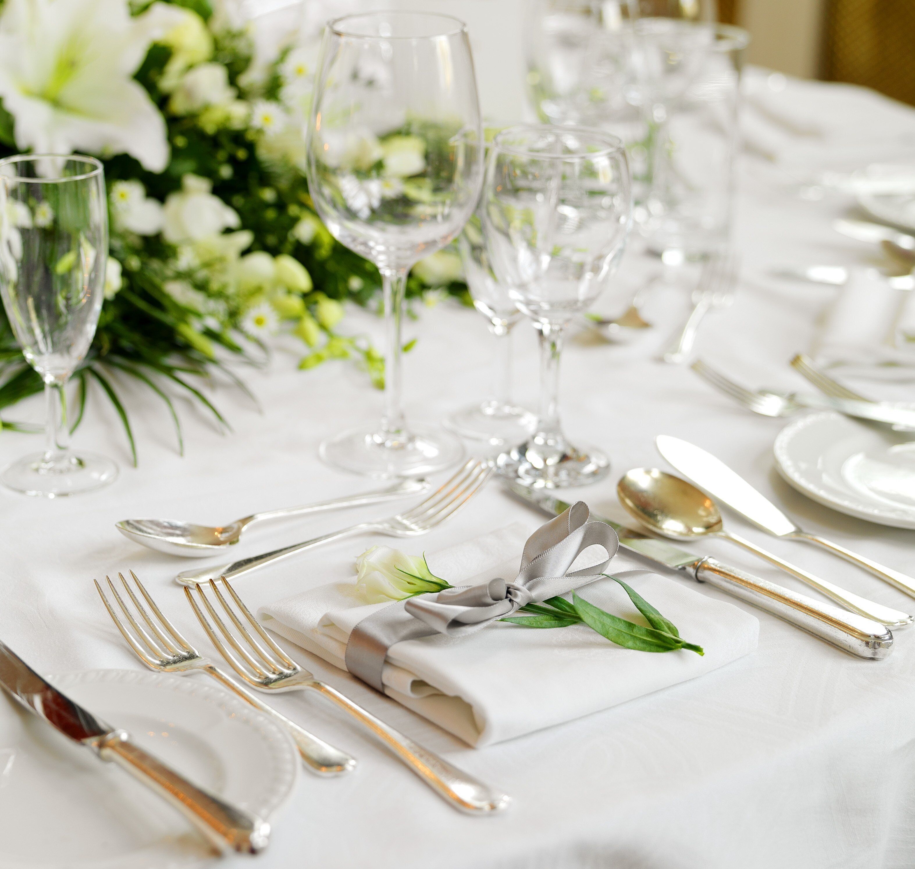 A table set with white tablecloth, silverware, and clear glassware, with a floral arrangement in the background.