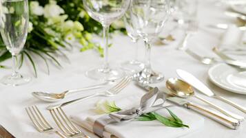 A table set with white tablecloth, silverware, and clear glassware, with a floral arrangement in the background.