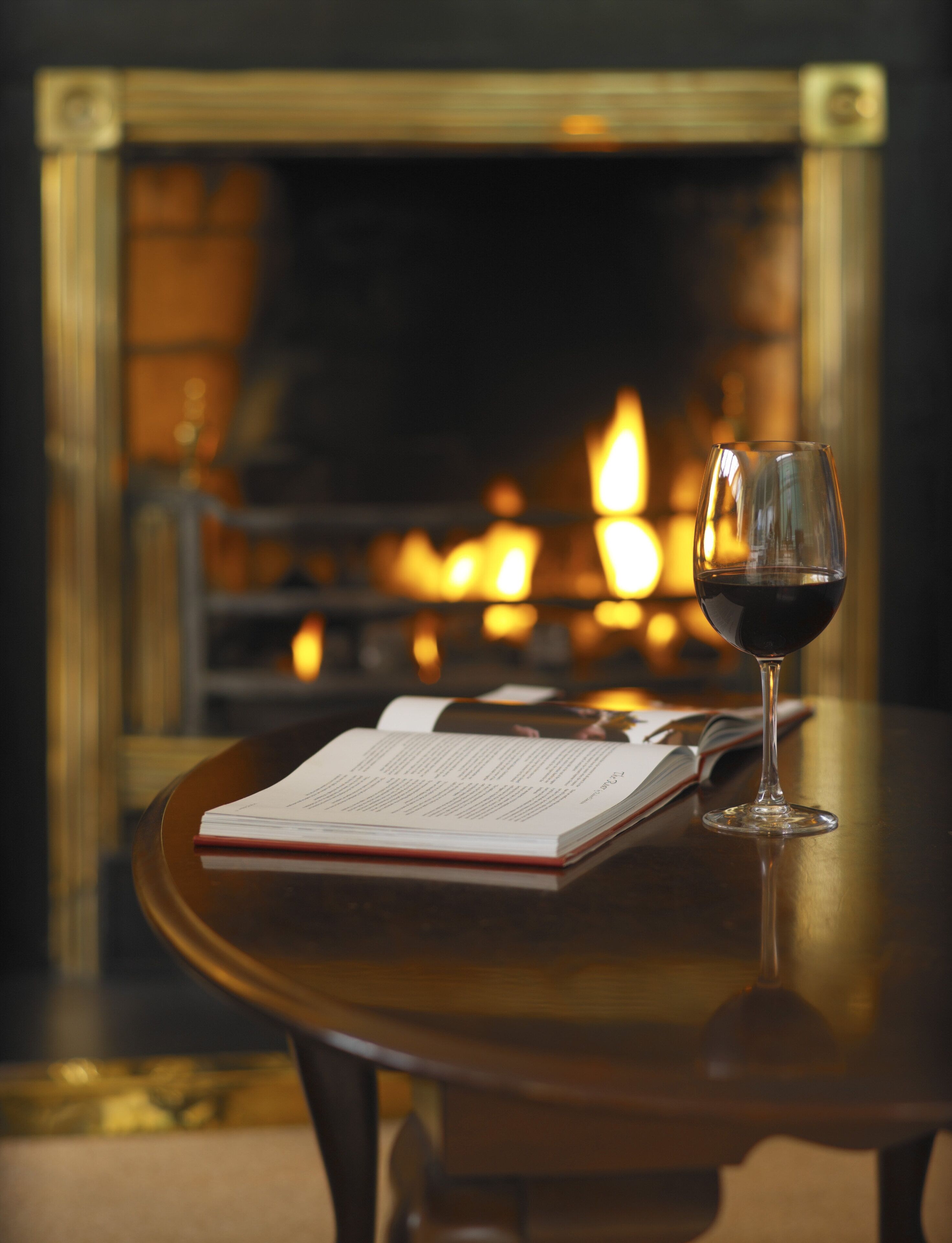 A book and a glass of red wine on a table with a fireplace in the background.