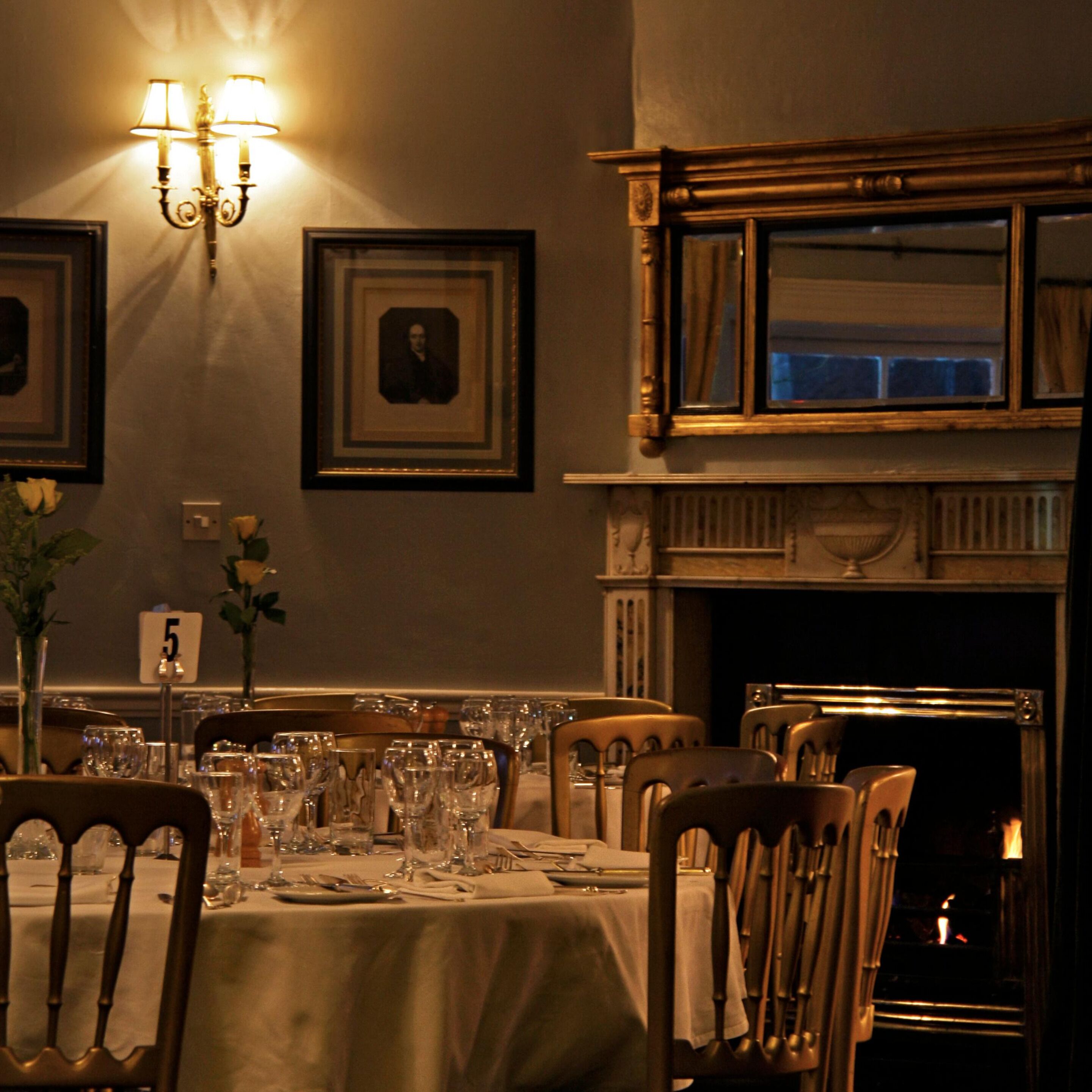 A dining area with a table set for four, chairs, a wall-mounted light fixture, framed pictures, and a fireplace.
