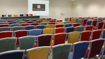 Un salón de conferencias con filas de sillas coloridas, una pantalla de proyector mostrando el logotipo del hotel y un cartel con el nombre de la sala.