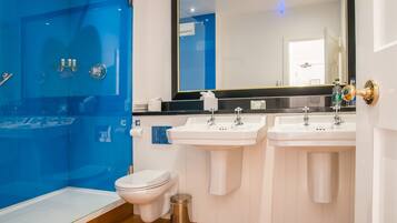 A modern bathroom with a glass shower enclosure, two sinks, a toilet, and a trash can.