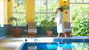 Zona de piscina con suelo de baldosas, una silla, una planta en maceta y una persona caminando.