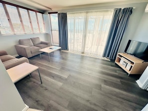 A modern living room with a sofa, coffee table, TV stand, and large windows with curtains.