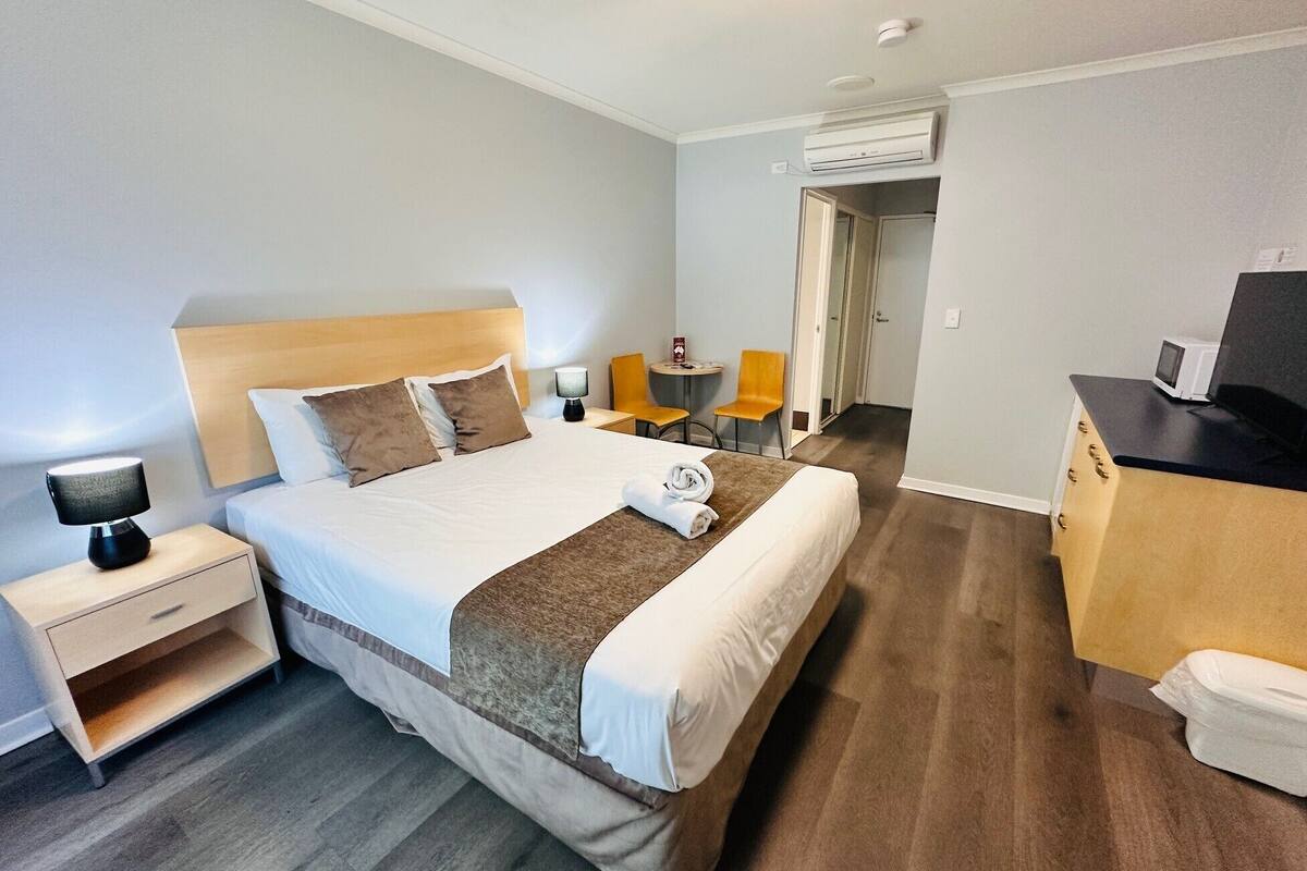 Una habitación de hotel con cama, mesita de noche, lámpara, silla, escritorio y un equipo de aire acondicionado.
