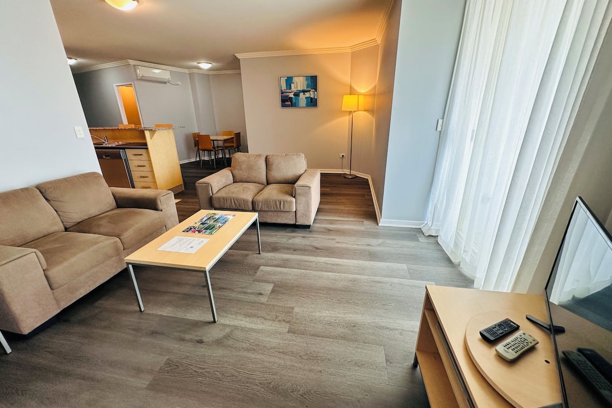 Una sala de estar con un sofá, una mesa de centro, un mueble para la televisión con mandos a distancia y una zona de comedor al fondo.