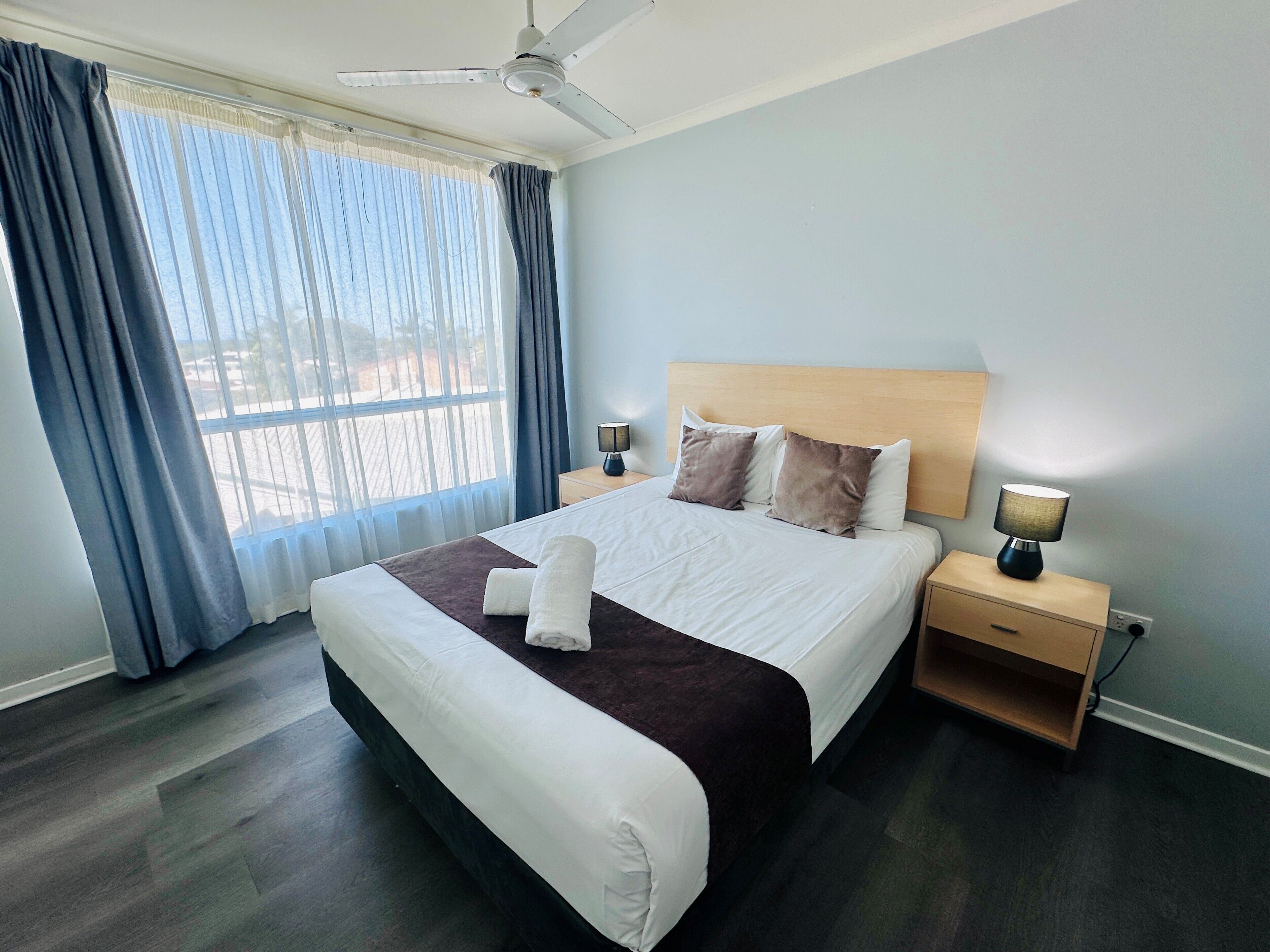 Una habitación de hotel con una cama, ventilador de techo, mesita de noche con lámpara y ventana con cortinas.
