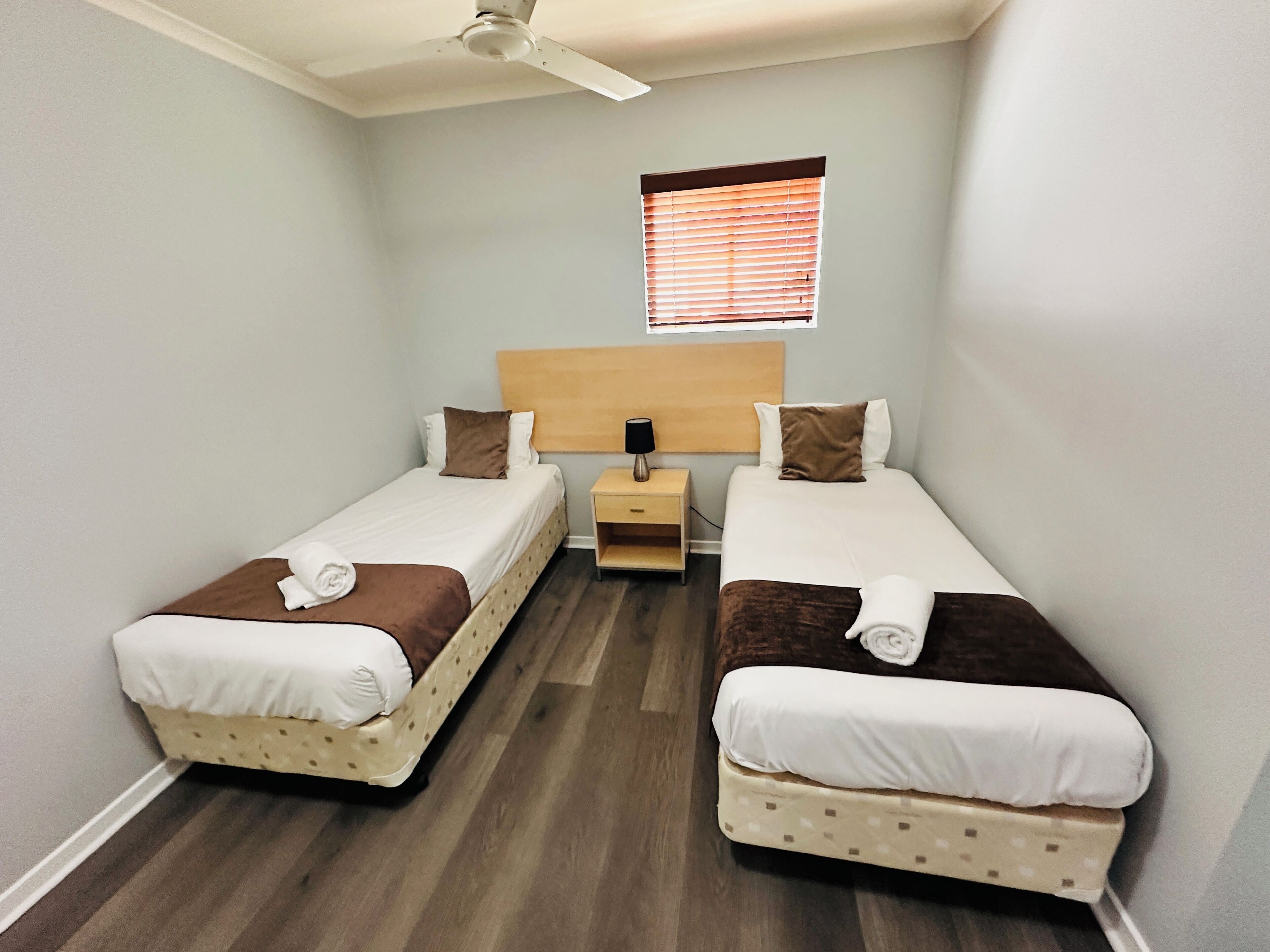 Une chambre double avec deux lits, une table de chevet, un ventilateur de plafond et une fenêtre avec des stores.