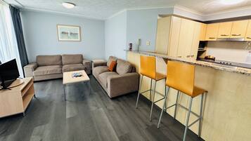 Una sala de estar moderna con un sofá, una mesa de centro y un mueble para la televisión. Incluye una zona de cocina con una encimera y taburetes de bar.