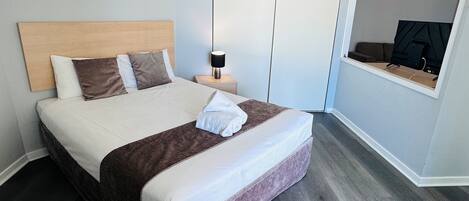 A neatly made bed with a headboard, a bedside table with a lamp, and a wall-mounted TV.